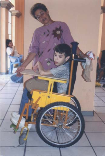 This child with floppy cerebral palsy has low muscle tone and slumps forward when he sits. The adjust-able table on a wheelchair designed at PROJIMO can be raised to lift his arms and shoulders enough so that he can hold up his head. As he gains strength, his mother can lower the table bit by bit.