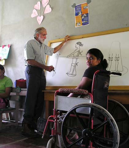 Esta joven en Honduras, cuyas piernas y caderas están paralizadas por la poliomielitis, usa una silla de ruedas, pero está muy ansiosa por caminar, y como sus brazos son muy fuertes, tiene buen potencial. Aquí, en un centro de rehabilitación de la aldea, David Werner dibuja un parapodio en la pizarra, que puede ser la mejor manera de comenzar a caminar.
